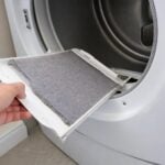 A hand pulling a lint screen coated in gray lint out of a home clothes dryer.