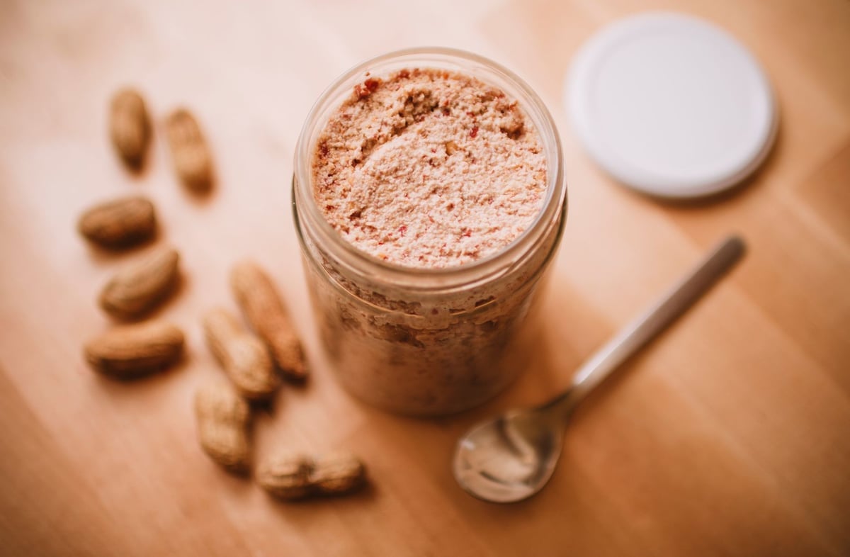 Delicious homemade peanut butter in a glass jar with peanuts and a spoon nearby.
