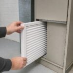 Hands sliding a new pleated air filter into a home furnace in a basement utility room.
