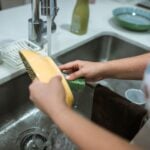 Hands washing a dish with a sponge at a kitchen sink.